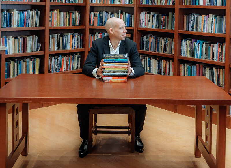 Man in a suit sits at a wooden table in a library, hands resting on a stack of colorful books against tall bookshelves.