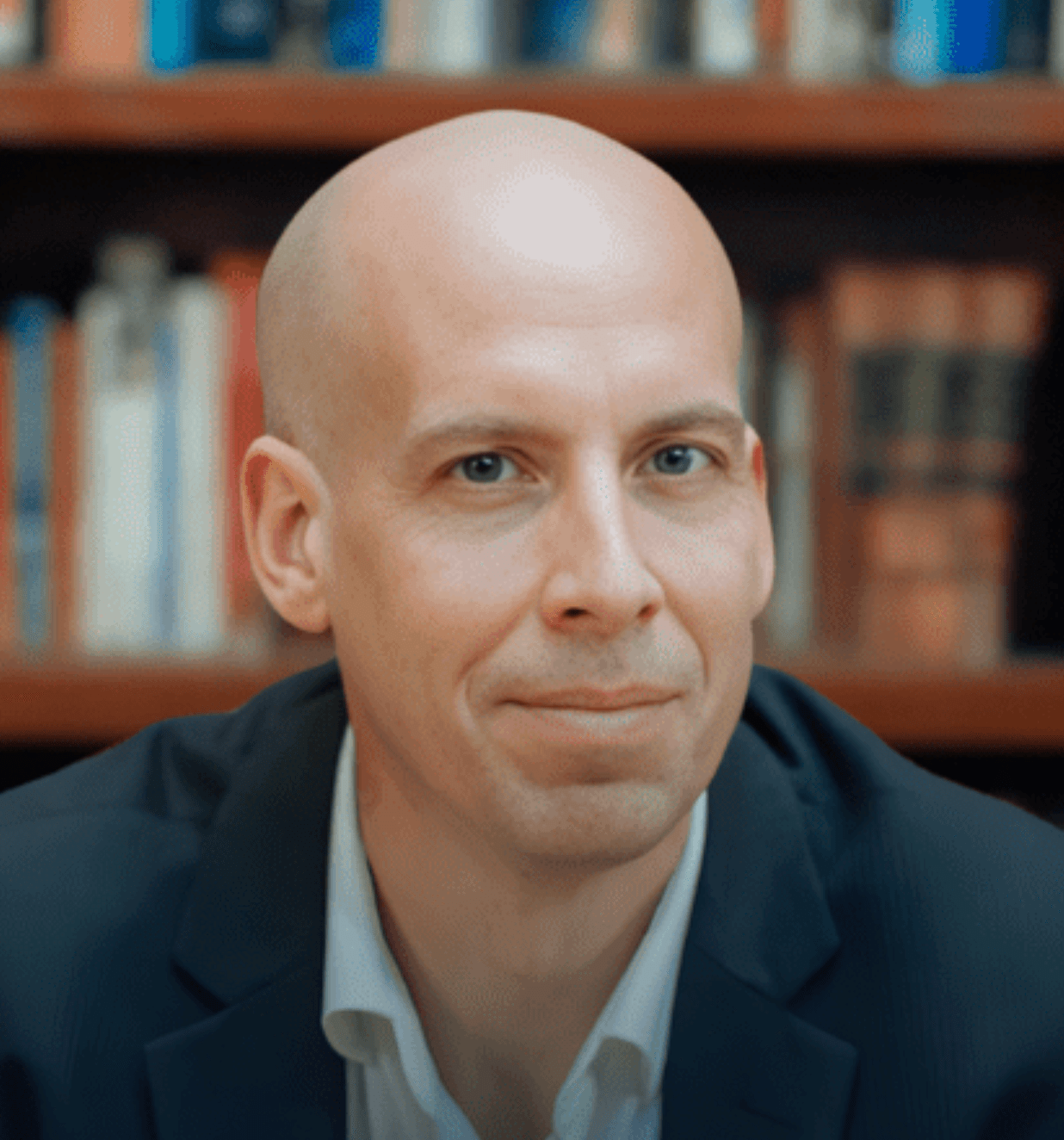 Man in a suit sits at a wooden table in a library, hands resting on a stack of colorful books against tall bookshelves.